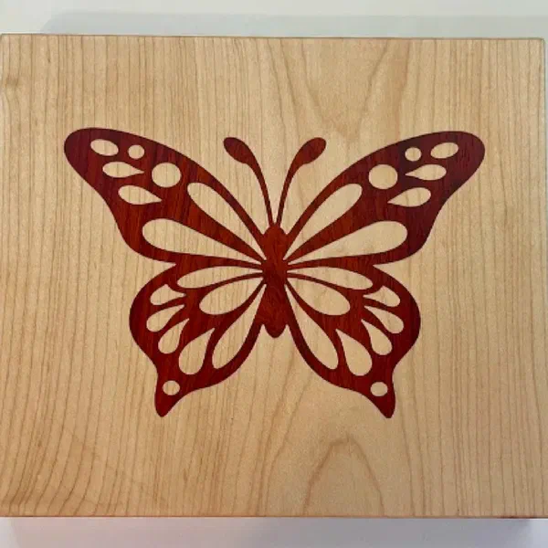 A red padauk butterfly inlaid flush into a maple panel, cut as a V-carve inlay.