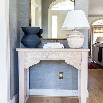 A pale carved console table against a wall with a lamp and vases on top.