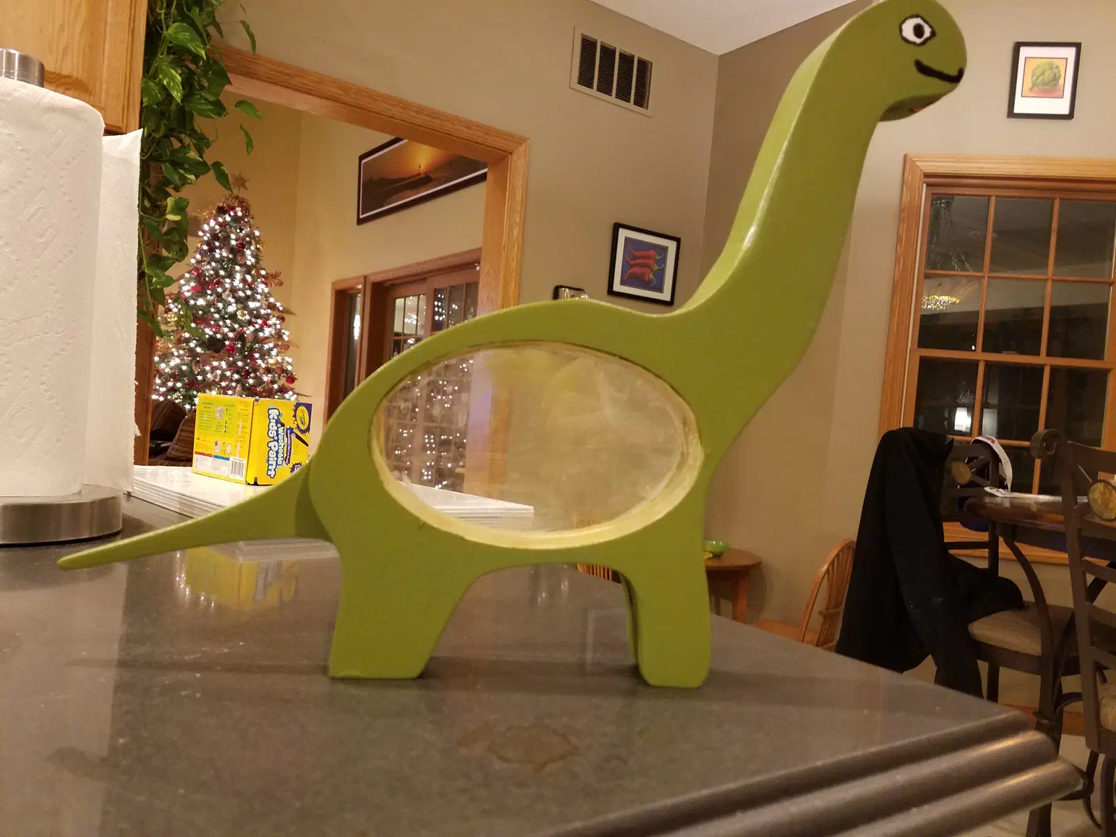 A painted green long-necked dinosaur coin bank with a clear oval belly standing on a kitchen counter
