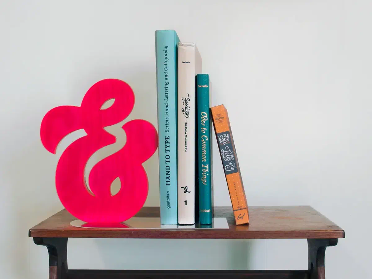 Whole pink acrylic-faced plywood ampersand on a clear base beside four books, against a plain pale wall.
