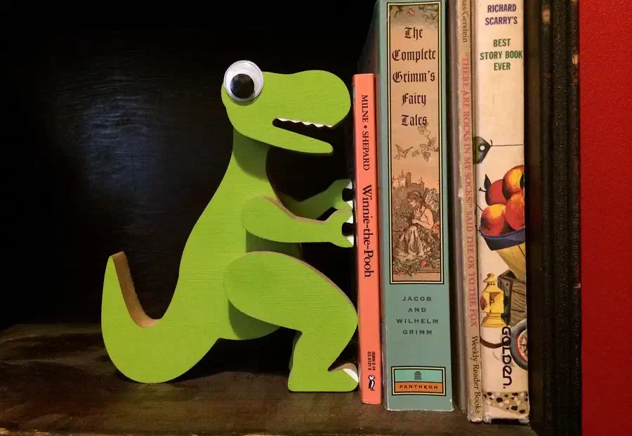 Whole green layered dinosaur bookend standing on a dark shelf beside books, with a large googly eye.