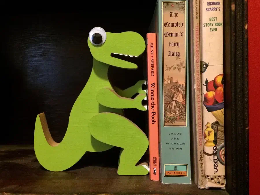 Whole green layered dinosaur bookend standing on a dark shelf beside books, with a large googly eye.