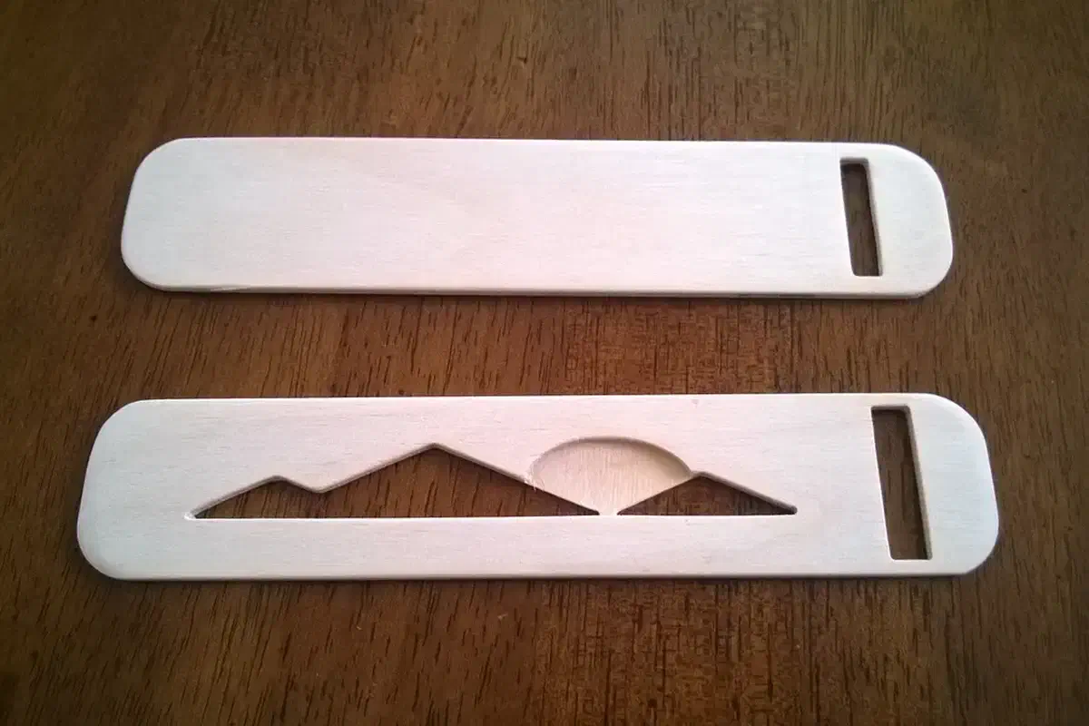 Two complete pale plywood bookmarks on a wooden table, one plain and one with a mountain-and-sun cutout, both with slots at the right end