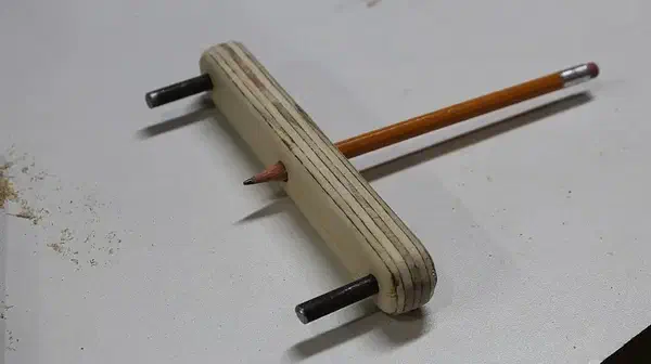 Finished layered plywood centerline tool with two metal pins and an orange pencil through its middle hole