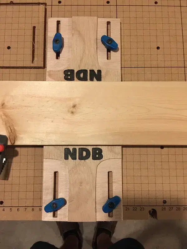 Two plywood side clamps with blue screws and NDB lettering gripping a wooden board