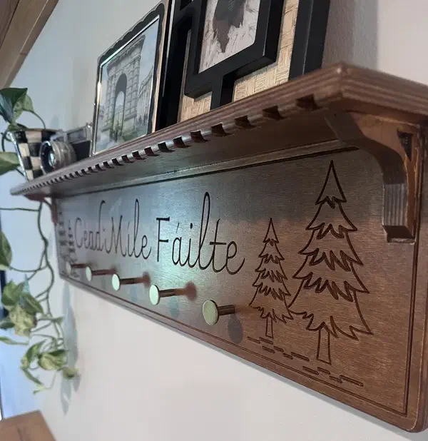 A finished wooden coat rack with pegs, carved Irish welcome lettering and trees, and a shelf holding framed pictures