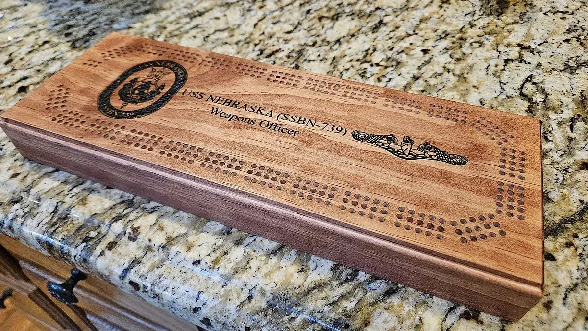 A rectangular wooden cribbage board with rounded scoring tracks and dark Navy insignia and lettering