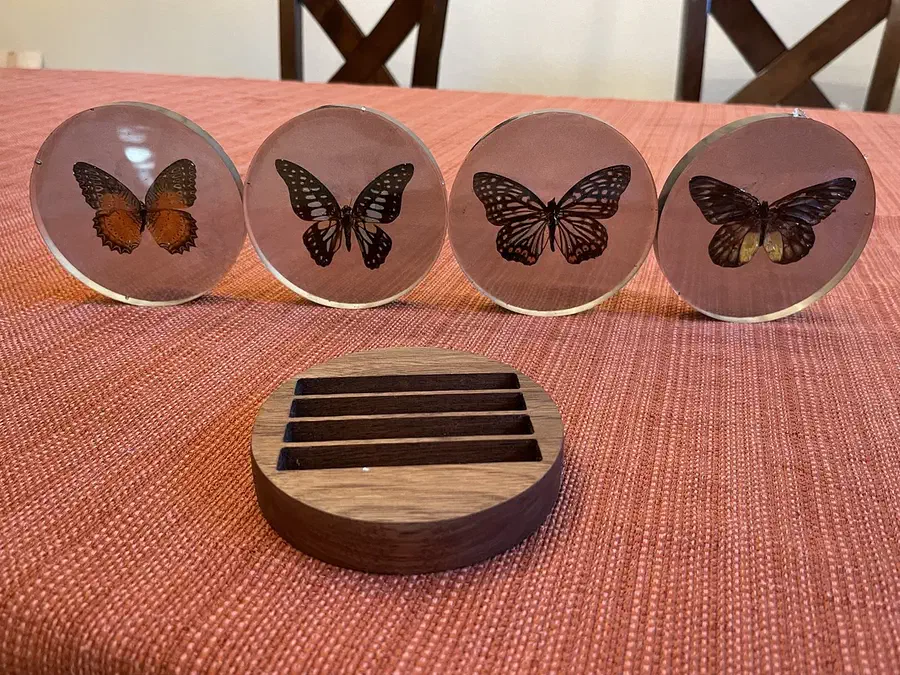 Round wooden slotted coaster holder in front of four clear butterfly resin coasters