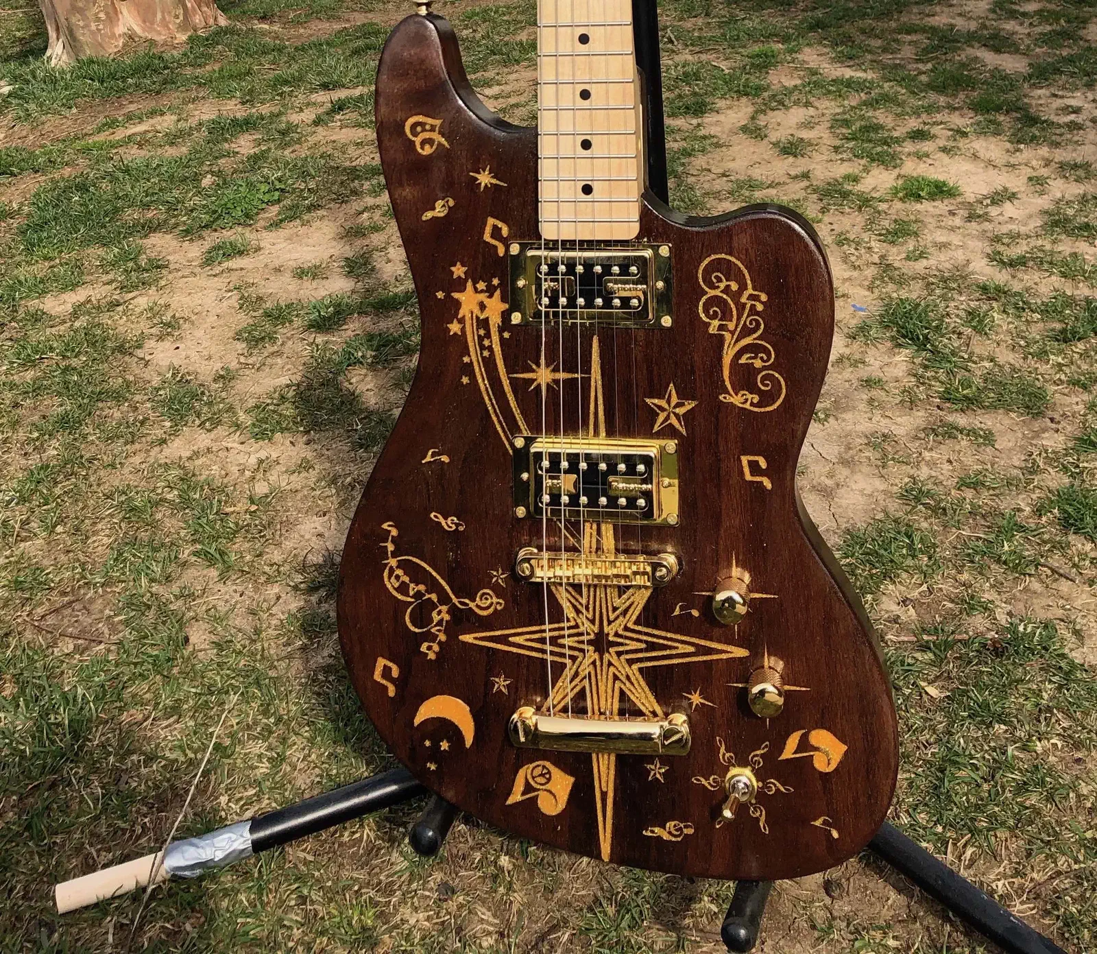 A walnut electric guitar body with gold-colored decorative patterns, dark pickups and metal hardware on an outdoor stand