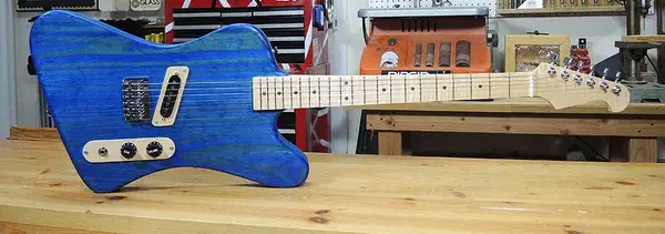 A blue electric guitar with a pale wooden neck resting horizontally on a workshop bench