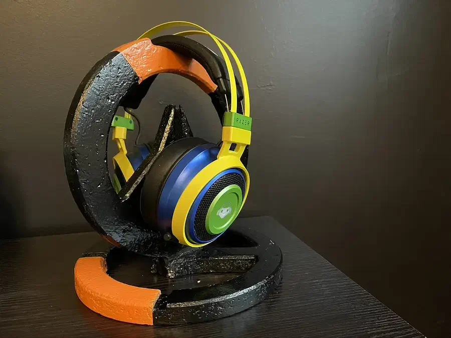 Blue, green and yellow headphones hanging on a black-and-orange Overwatch emblem stand with a matching circular base