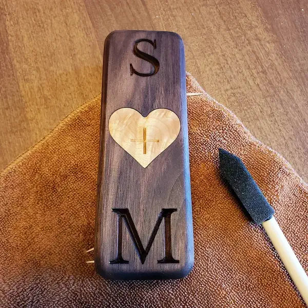 Long dark wooden necklace box with carved initials and a pale heart-shaped inlay on a towel