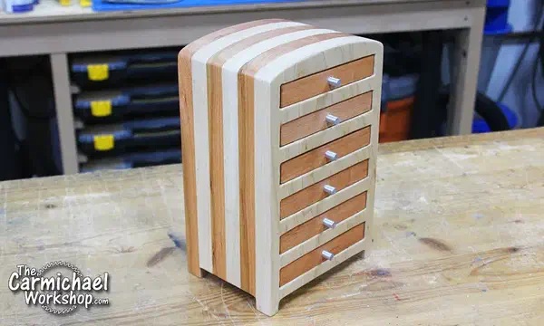 Six-drawer maple and cherry jewelry box with striped sides and aluminum pulls on a workbench