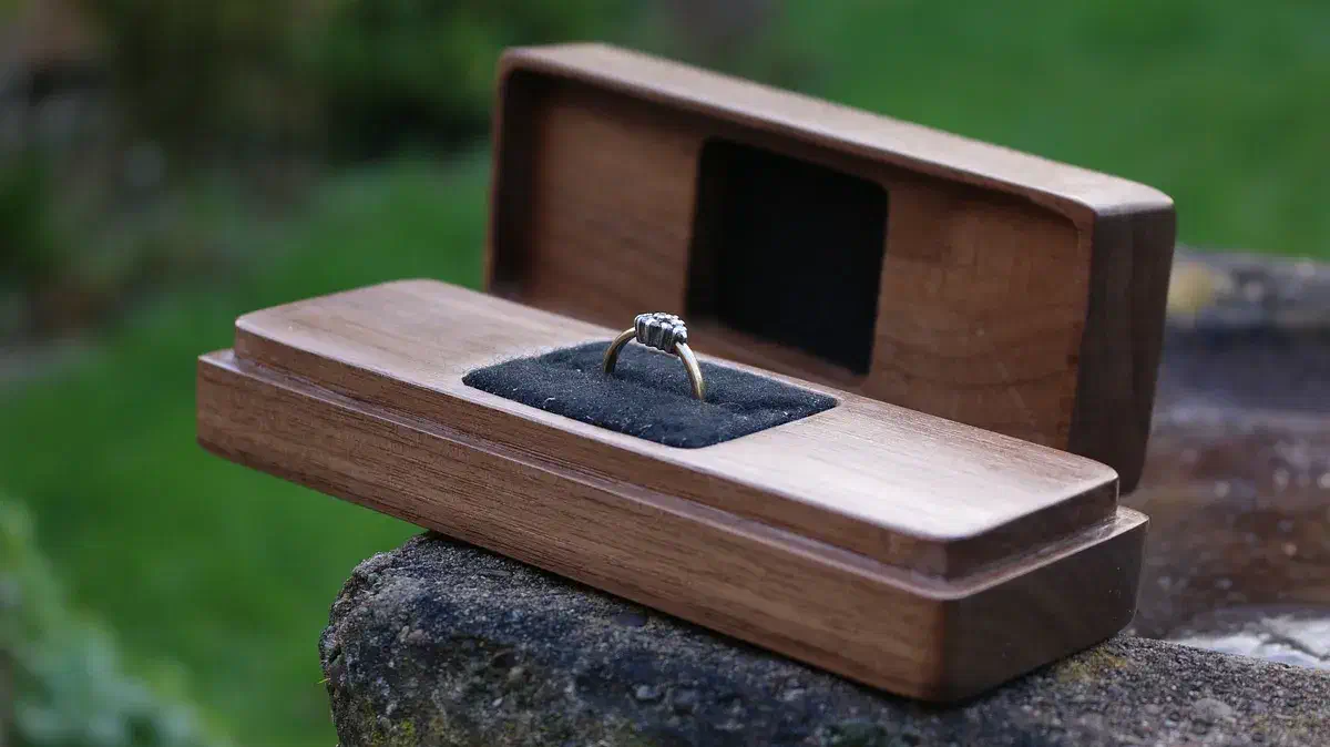 Long walnut ring box with its lid propped behind a ring held in black lining on a stone wall