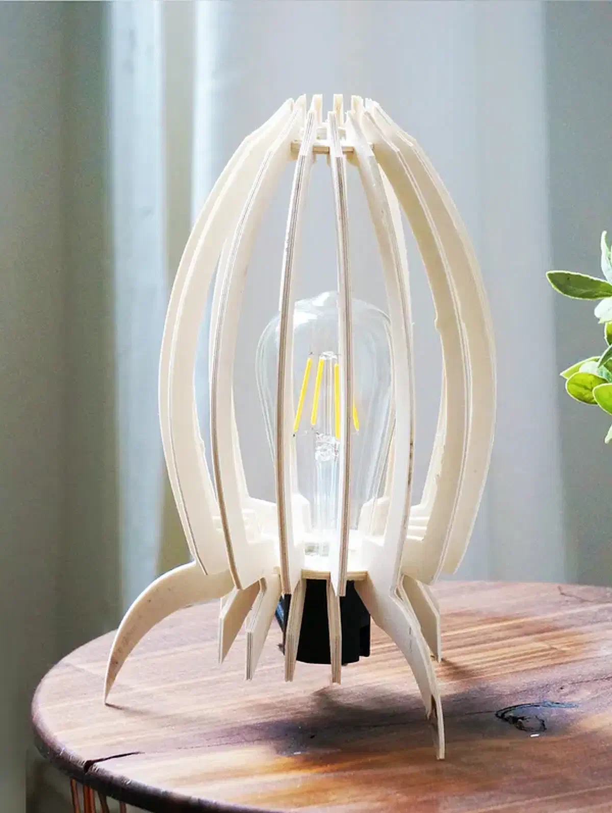 A pale plywood ribbed table lamp with pointed feet beside a potted plant