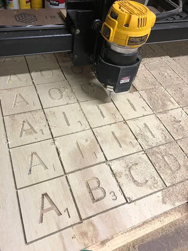 An X-Carve router carving large letter-and-point-value tiles in a plywood panel