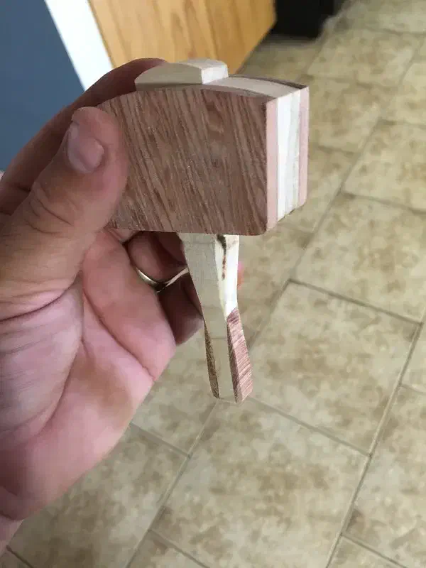 A hand holding a miniature wooden mallet with a pale square face and laminated handle