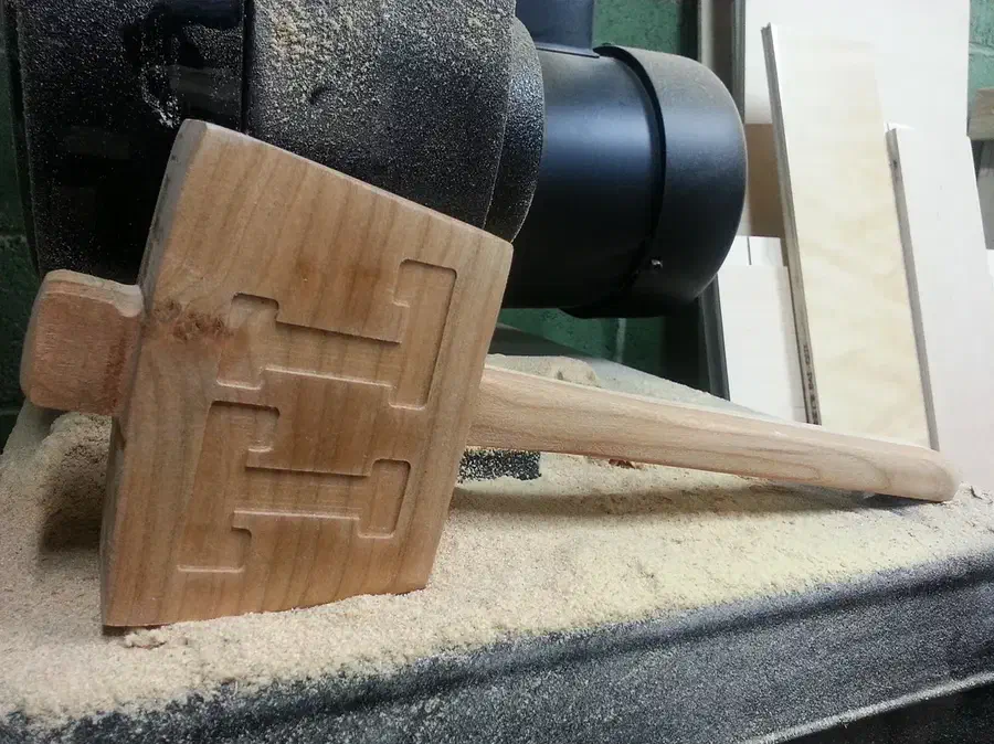 A laminated wooden mallet with carved initials on its broad head, resting beside a workshop sander