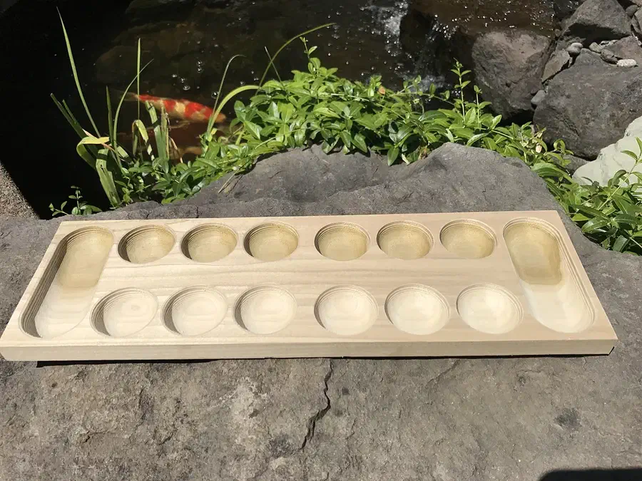 Light-colored Mancala board with twelve round bowls and two long end stores, with visible spiral tool marks, resting on rocks beside a pond