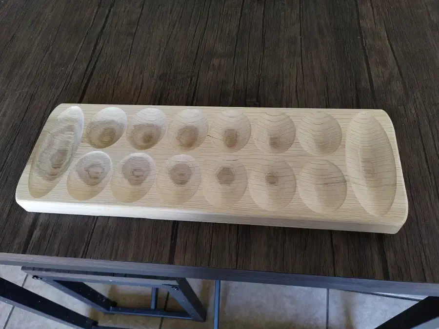 Finished pale wooden Mancala board with twelve oval bowl-shaped pits, two long end stores, and rounded outside corners on a dark table