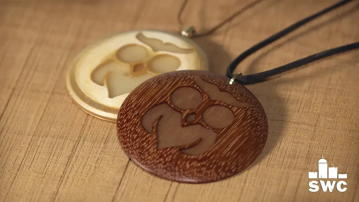 Two round wooden owl pendants, one pale and one reddish, with a black cord on a workbench