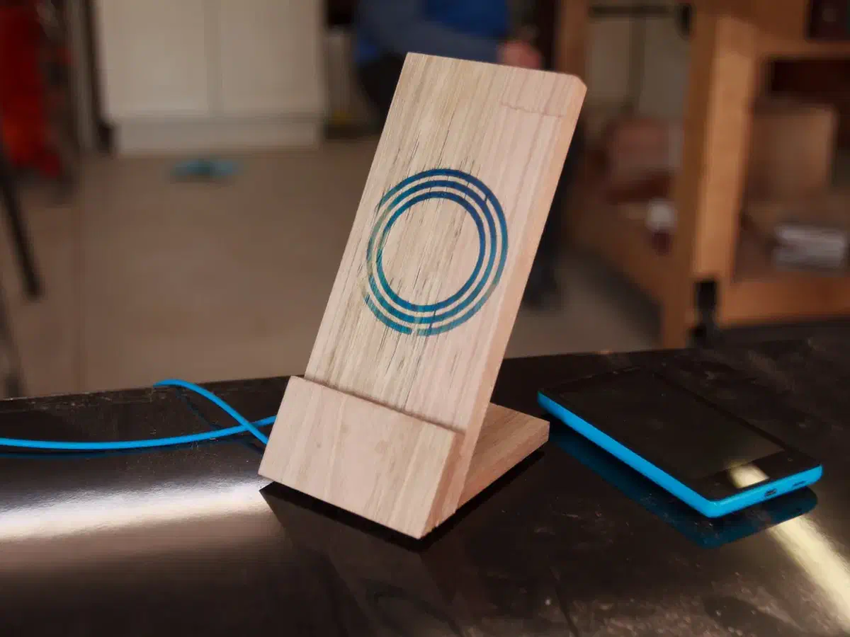 Wooden upright charging stand with blue carved rings, shelf and base beside a phone