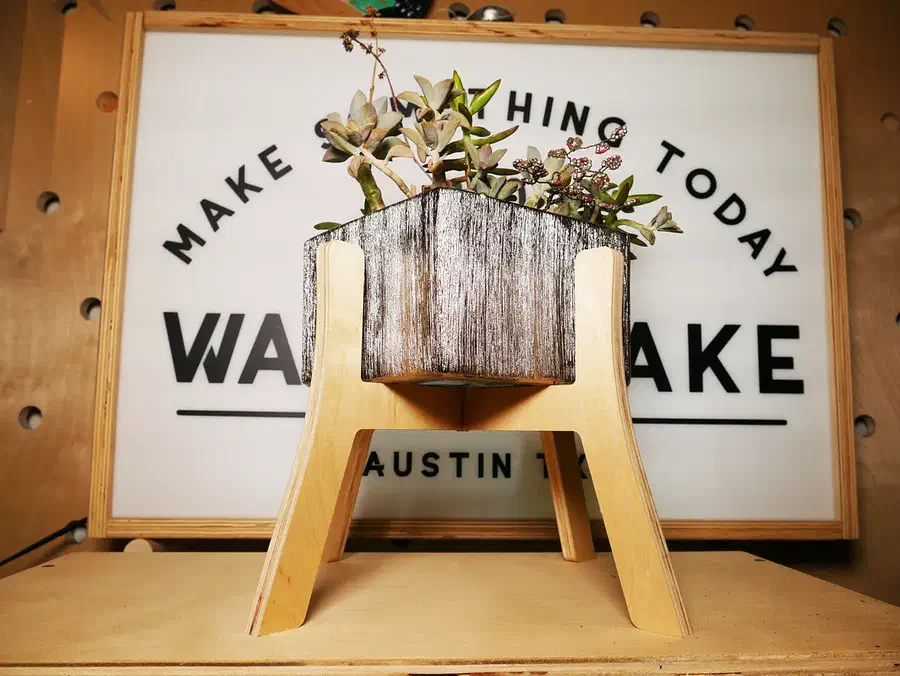 A wooden planter on a pale plywood stand with splayed legs in front of a workshop sign