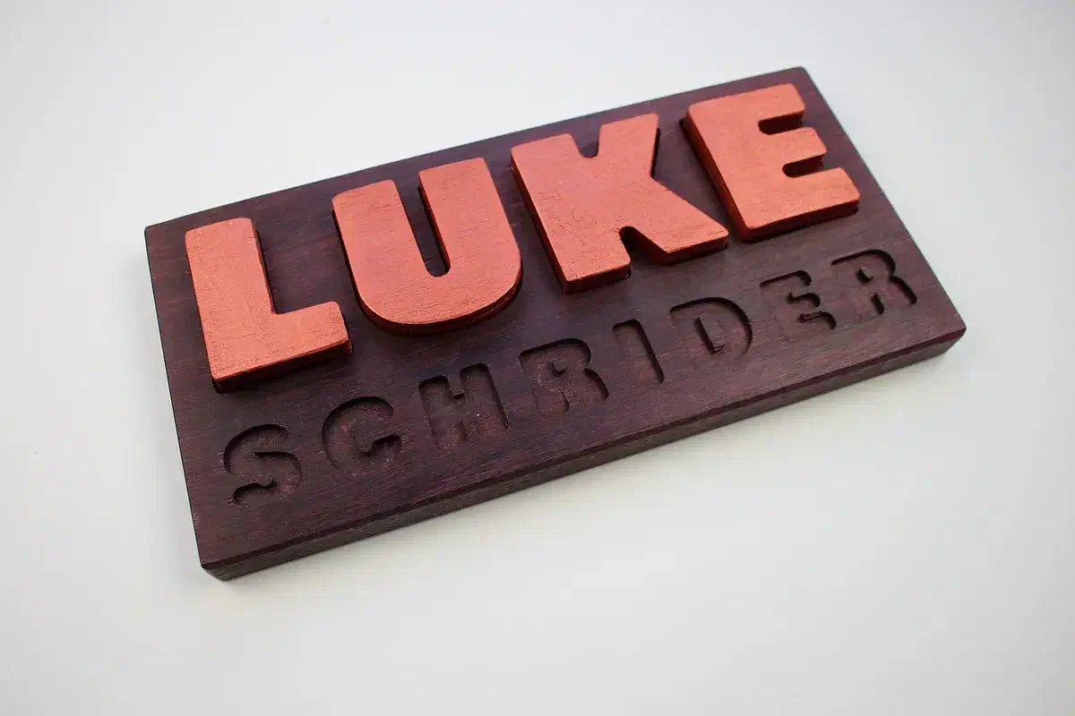 Removable coral-colored LUKE letters on a dark wooden puzzle board with a carved surname