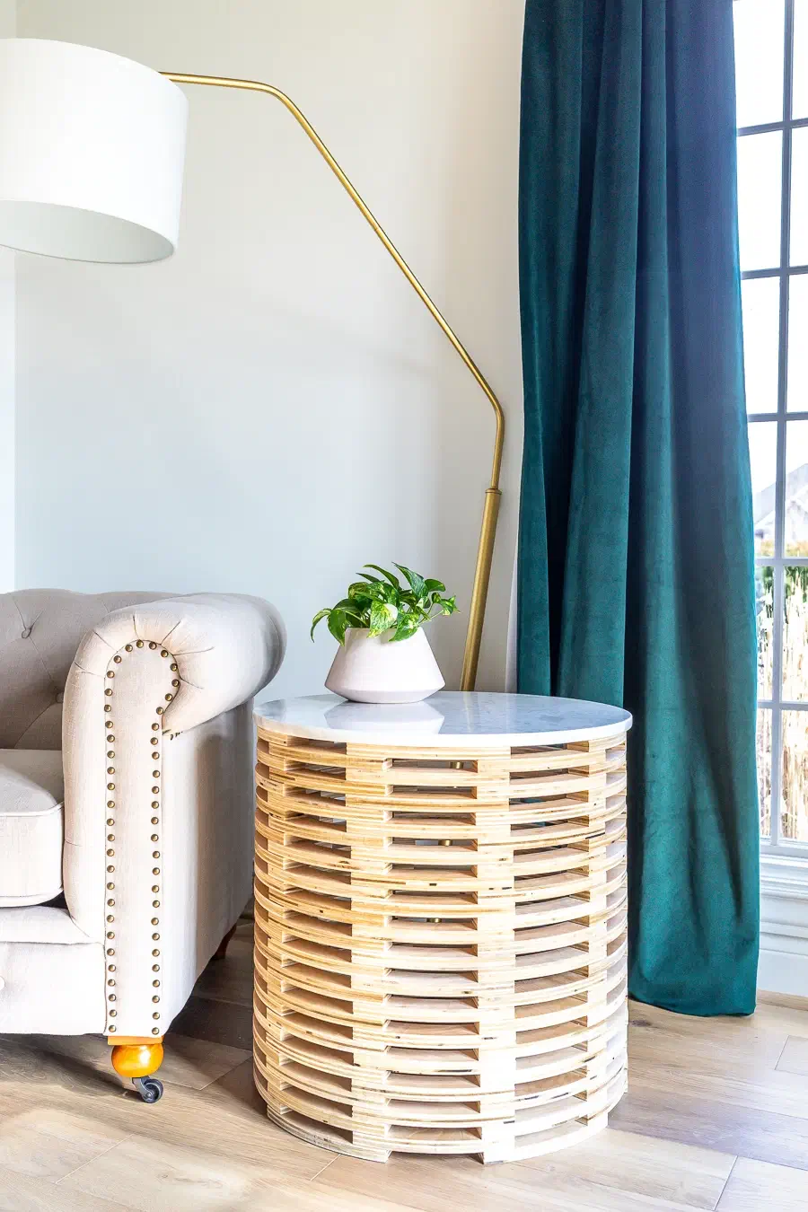 A cylindrical table built from stacked pale plywood slats with a white round top beside an upholstered armchair