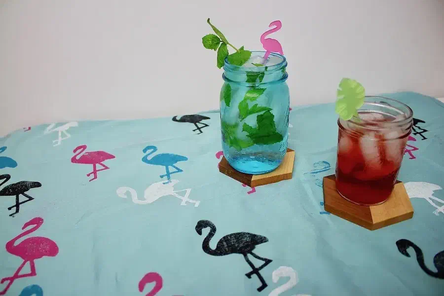A pale tea towel printed with flamingos, leaves and pineapples beneath two colorful drinks