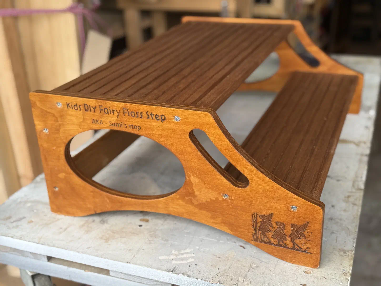 A wide two-step wooden stool with grooved brown treads and decorated side panels