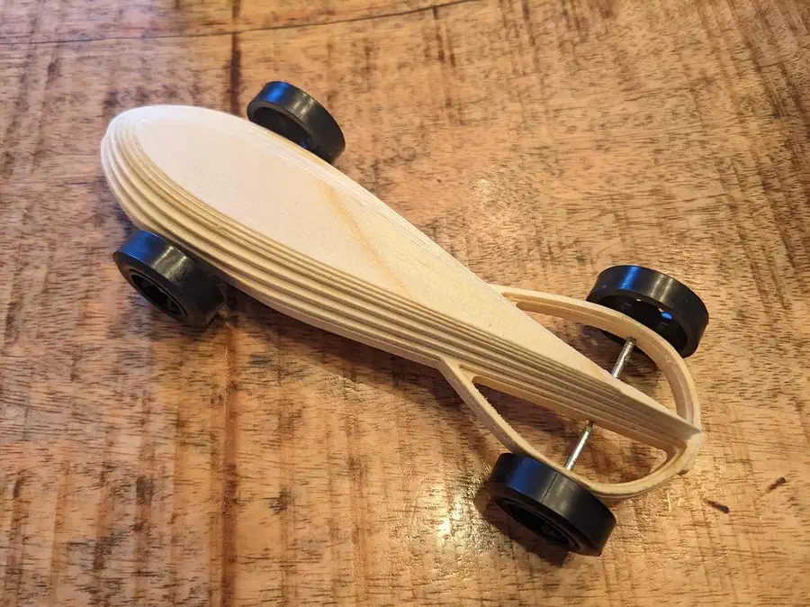 A narrow pale wooden pinewood derby car with black kit wheels on a workbench
