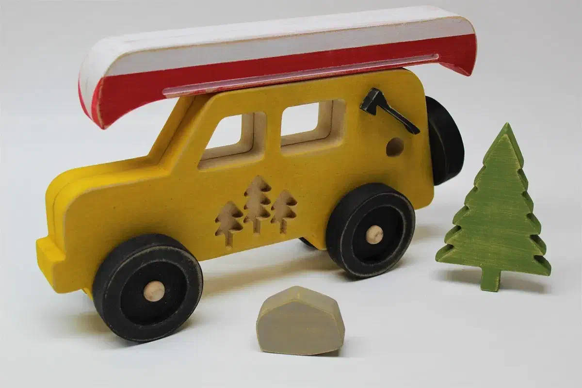 A yellow wooden car carrying a red and white canoe beside a small green wooden tree