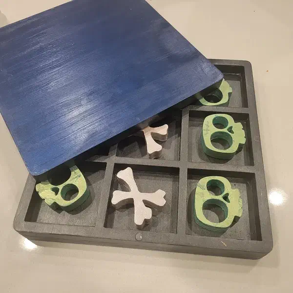 A painted wooden tic-tac-toe game with green skull pieces, pale crossbones and a dark lift-off lid