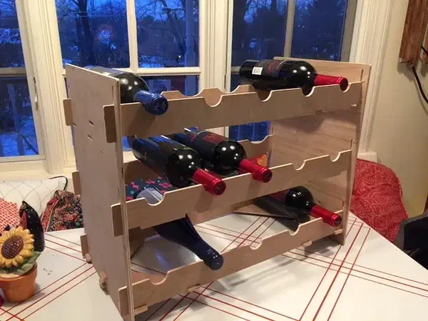 A plywood wine rack with three rows of bottle supports standing on a table beside a window