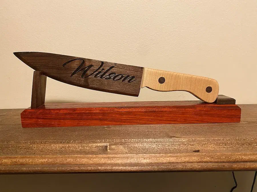 A wooden cake knife with a dark engraved blade and pale handle displayed on a wooden stand